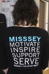 Mentorship and Training Coordinator Liz Longfellow at MISSSEY displaying the organization's staff shirts.